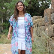 Woman wearing a colorful tunic and white dress standing in front of a stone wall with greenery.